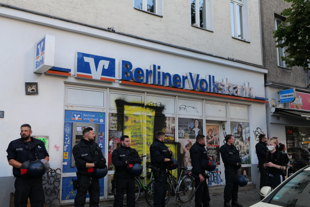 Eine Gruppe von Polizisten steht vor einer Berliner Volksbank, einige halten Helme in den Händen, rechts ein Auto und links ein Baum; im Hintergrund sind Gebäude mit Fenstern und Texttafeln zu sehen.