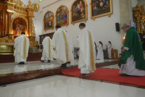Eine Gruppe von Priestern geht einen roten Teppich in einer Kirche entlang, mit Menschen, die auf dem Boden vor ihnen und auf der rechten Seite stehen, und Foto-Rahmen an der Wand im Hintergrund.