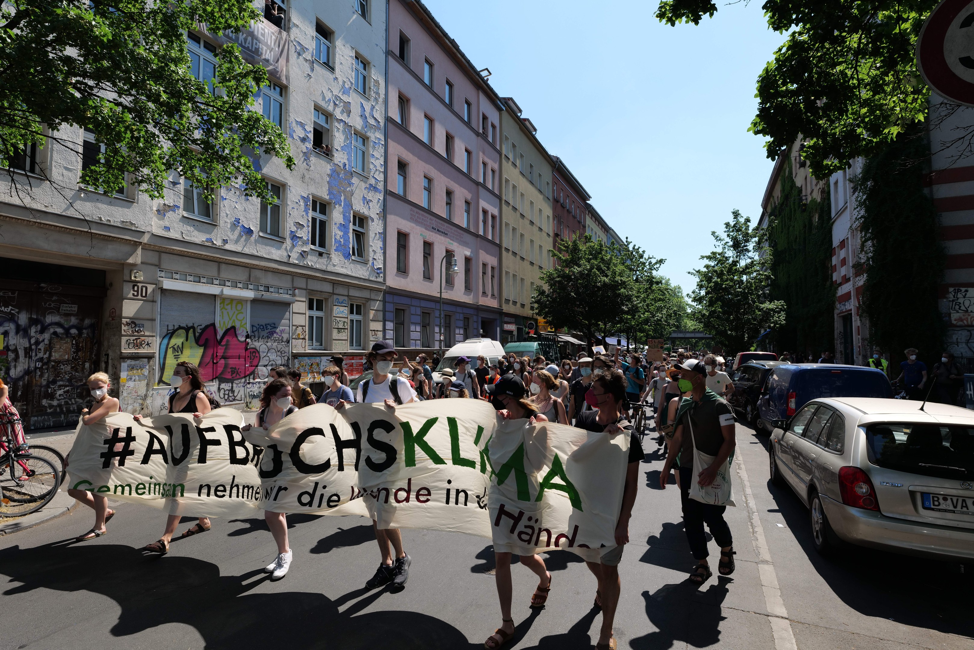 Eine Gruppe von Menschen marschiert mit einem Transparent die Straße entlang, mit parkenden Autos auf der rechten Seite, einem Radfahrer auf der linken Seite und Gebäuden, Bäumen und einem Schild im Hintergrund unter einem sichtbaren Himmel.