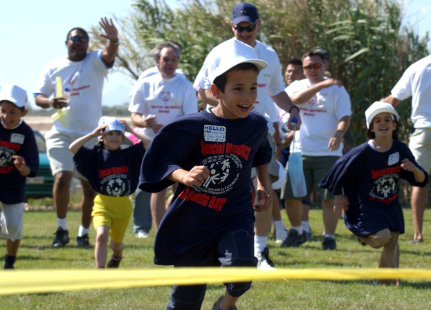 Gruppe von Kindern, die auf einem Rasenfeld in einem Rennen laufen, mit einem gelben Band im Vordergrund, einige tragen Kappen und Schutzbrillen, andere halten Gegenstände, Bäume und klarer blauer Himmel im Hintergrund.
