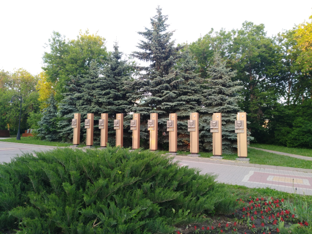Ein Park mit einer Reihe von Bäumen in der Mitte, umgeben von Blumenpflanzen, Gras, einem Weg, einem Straßenpfahl, einer Gruppe von Bäumen und einem bewölkten Himmel, mit einem Schild, das an die Opfer der Tschernobyl-Katastrophe erinnert.