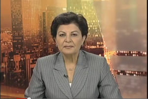 Eine Frau in einem Business-Outfit sitzt vor einem Fernseher, mit ein paar Gebäuden im Hintergrund, die als Nachrichtensprecherin mit Text am unteren Bildschirmrand erscheint.