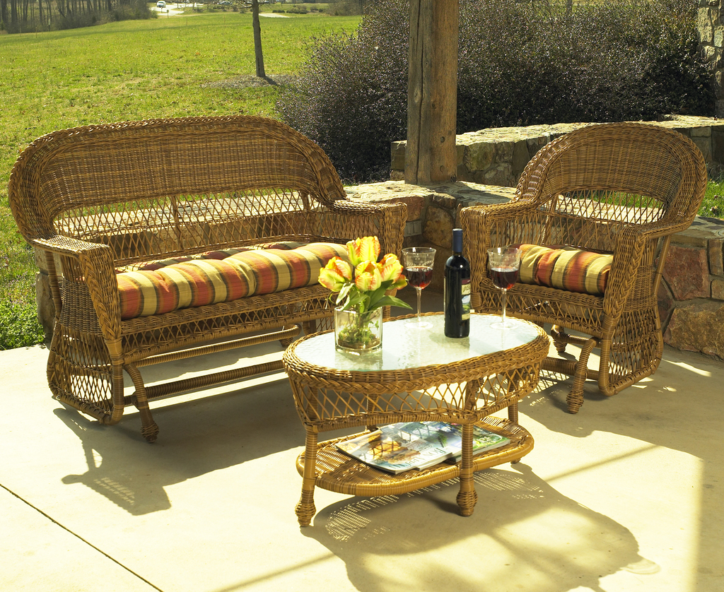 Eine Zweiersofa und ein Einzelsofa mit einem Tisch dazwischen, der eine Weinflasche, zwei Gläser und eine Vase mit gelben Blumen sowie Magazine darunter enthält, vor einer Felswand mit Büschen, Gras und Bäumen im Hintergrund.