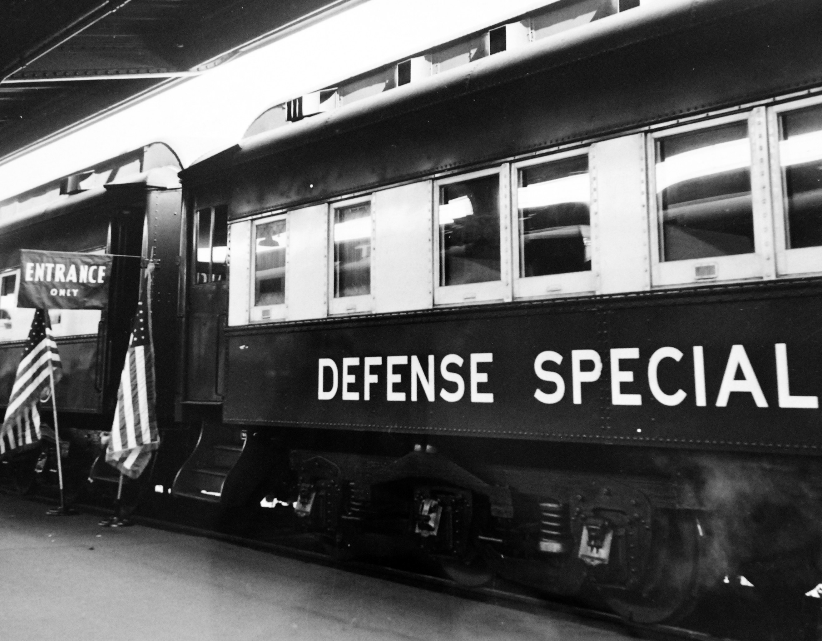 Schwarz-weißes Foto eines Zugs mit der Aufschrift "Defense Special", der auf Gleisen geparkt ist, mit zwei Fahnen links und einer Decke oben.