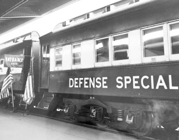 Schwarz-weißes Foto eines Zugs mit der Aufschrift "Defense Special", der auf Gleisen geparkt ist, mit zwei Fahnen links und einer Decke oben.