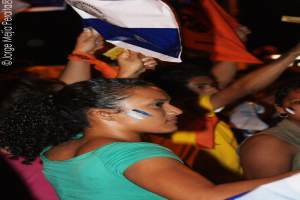 Eine Frau in einem grünen T-Shirt steht auf einer Demo und hält ein weißes Schild in der Hand, während viele andere Menschen hinter ihr stehen.