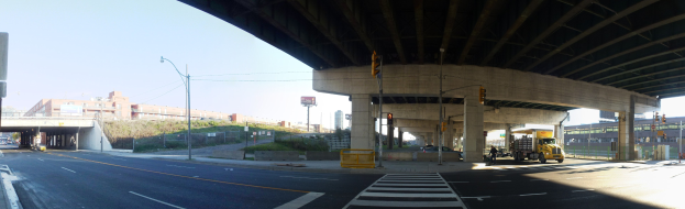 Lastwagen unter einer Brücke auf einer Stadtstraße mit Gebäuden, Straßeninfrastruktur, Fahrzeugen, Grünflächen und einem klaren Himmel.