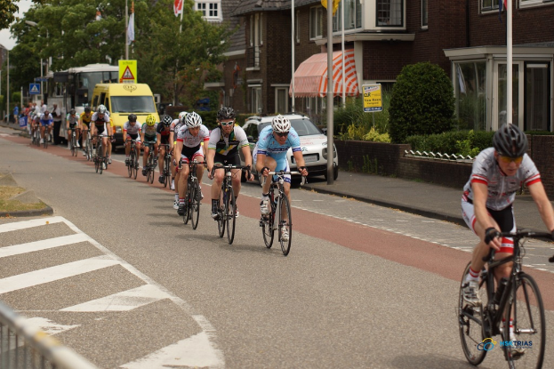 Menschen sitzen auf Fahrrädern mit Helmen auf einer Straße mit parkenden Autos, Fahnen an Masten und Gebäuden mit Bäumen im Hintergrund.