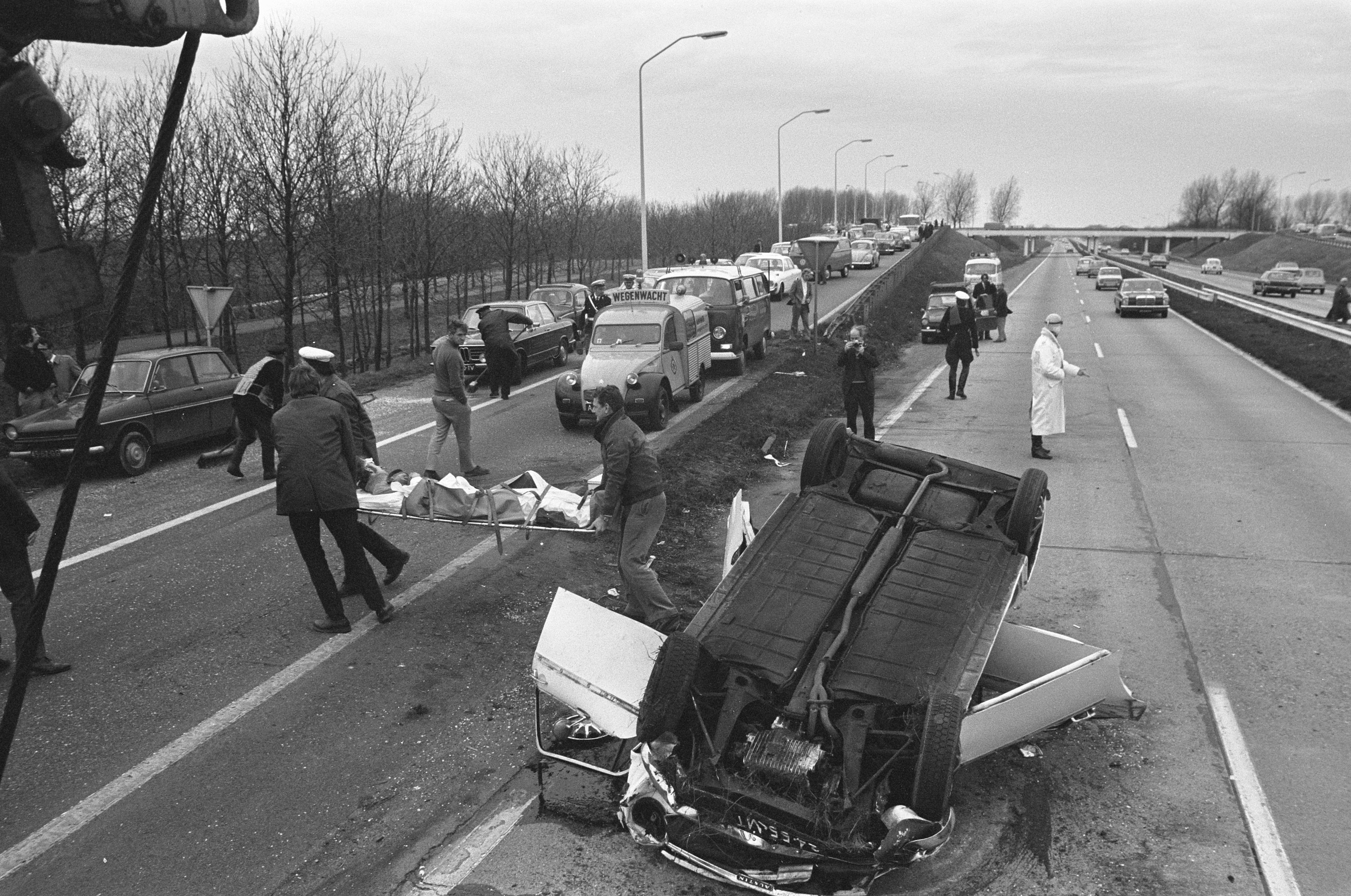 Schwarzes und weißes Foto eines umgestürzten Autos am Straßenrand mit beschädigten Fahrzeugen in der Nähe und einer Gruppe von Menschen drumherum, mit Laternenmasten, Bäumen, einer Brücke und Himmel im Hintergrund.