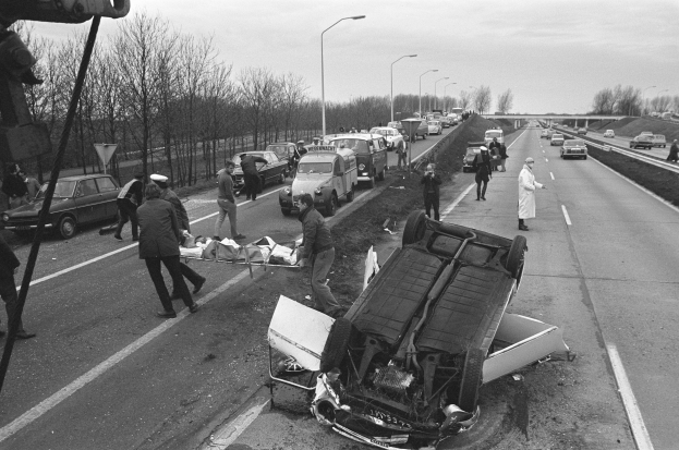 Schwarzes und weißes Foto eines umgestürzten Autos am Straßenrand mit beschädigten Fahrzeugen in der Nähe und einer Gruppe von Menschen drumherum, mit Laternenmasten, Bäumen, einer Brücke und Himmel im Hintergrund.