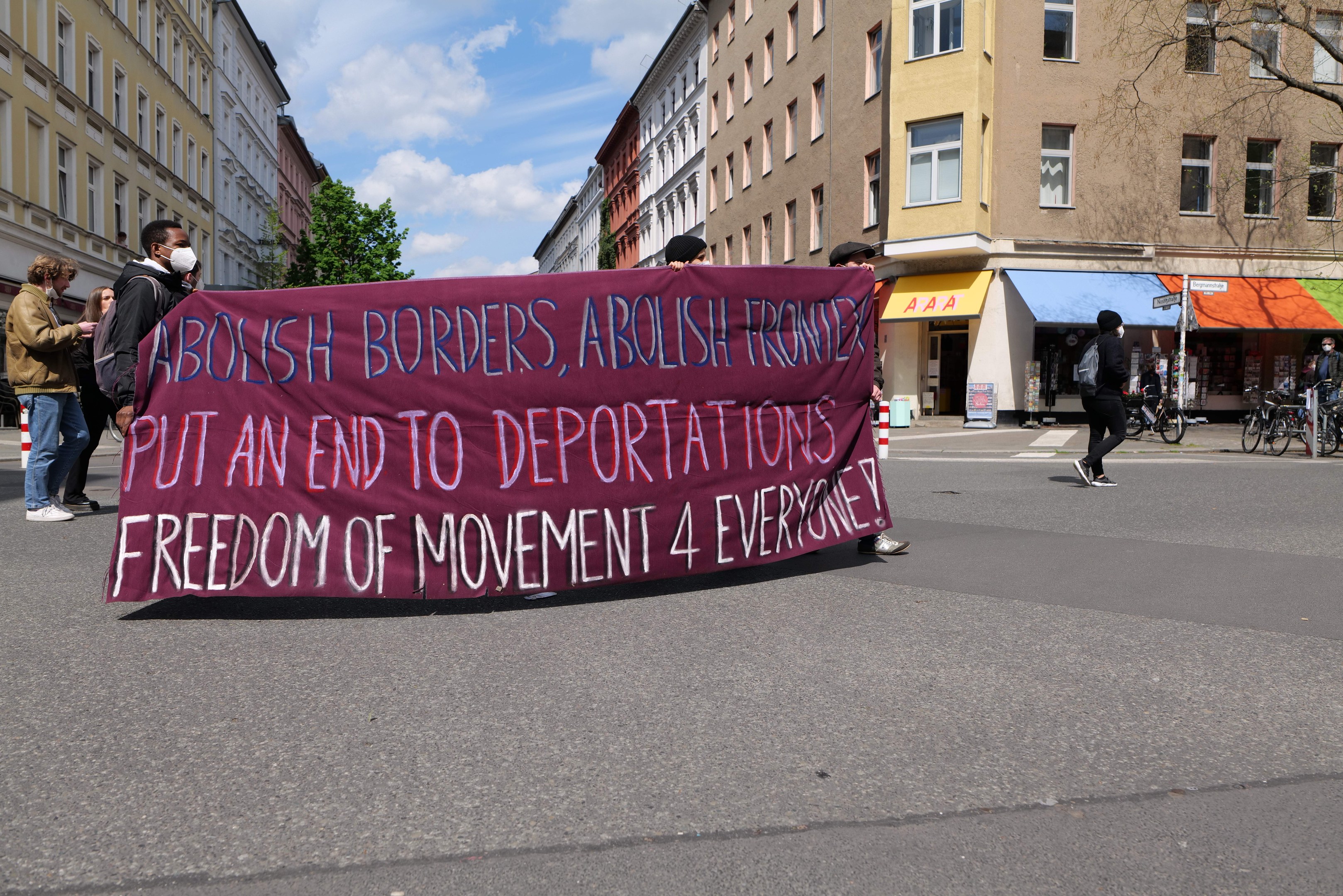 Eine Gruppe von Menschen marschiert mit einer Fahne, auf der "Offene Grenzen, Ende der Abschiebungen" steht, durch eine Stadtstraße, flankiert von Gebäuden, Bäumen und Fahrrädern bei bewölktem Himmel.
