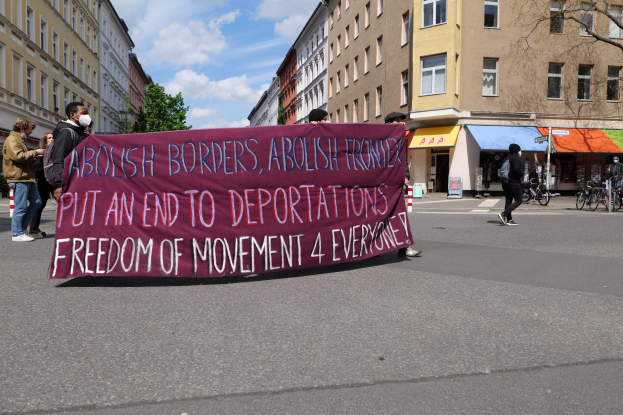 Eine Gruppe von Menschen marschiert mit einer Fahne, auf der "Offene Grenzen, Ende der Abschiebungen" steht, durch eine Stadtstraße, flankiert von Gebäuden, Bäumen und Fahrrädern bei bewölktem Himmel.