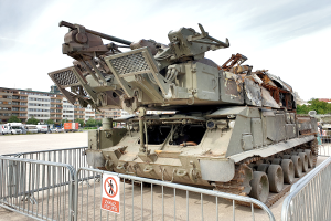 Ein gepanzerter Mannschaftstransporter vor einem Metallzaun mit einem daran befestigten Schild, mit Fahrzeugen, Bäumen, Gebäuden und einem klaren blauen Himmel im Hintergrund.