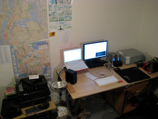 Ein Raum mit einem Tisch, auf dem ein Laptop, ein Lautsprecher, ein Monitor, eine Tastatur, ein Ball, eine Maus, ein Scanner und Papiere liegen, und ein weiterer Tisch mit Elektronikgeräten, mit Charts und Postern an der Wand dahinter.