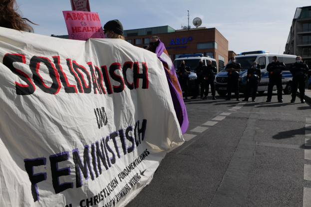 Eine Gruppe von Menschen marschiert auf einer Straße, trägt eine Fahne mit den Worten "Solidarität und Feminismus", mit parkenden Fahrzeugen und Gebäuden im Hintergrund unter einem klaren blauen Himmel.