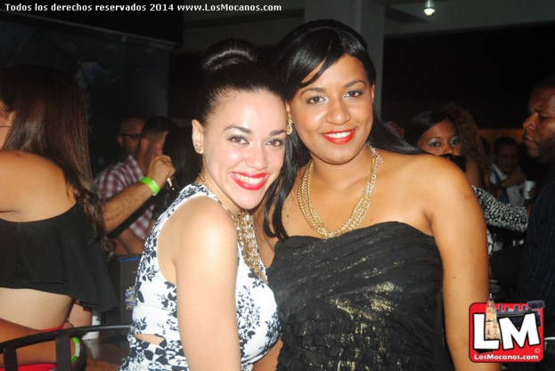 Zwei Frauen in modischer Kleidung stehen und lächeln in einer Bar, während im Hintergrund andere Leute tanzen und eine Lampe an der Decke leuchtet.