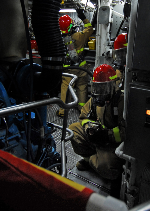 Feuerwehrleute in Helmen und Schutzausrüstung arbeiten an einem Feuerlöschwagen mit Rohren und einer Lampe im Hintergrund.