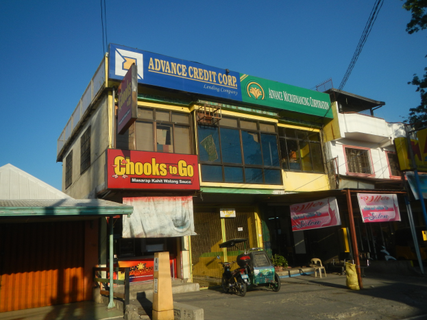 Ein Gebäude mit einer Tafel, auf der 'Advance Credit Corp' steht, umgeben von Fahrzeugen, Werbetafeln, Strommasten, Kabeln, Bäumen und einem klaren blauen Himmel.