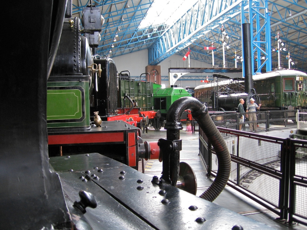 Mehrere Zugmaschinen stehen in einem Abstellgleis mit zahlreichen Eisenstangen darüber. Rechts verläuft ein Zaun entlang einer Bahnstrecke mit einem Zug. Zwei Personen stehen in der Mitte der Szene nahe dem Zaun.