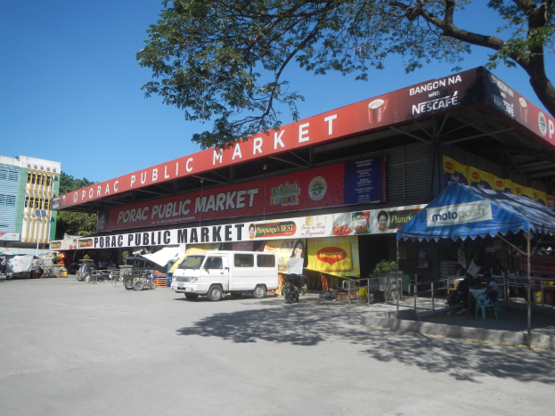 Ein ├Âffentlicher Markt mit einem vor ihm geparkten Van, umgeben von Geb├Ąuden, Gesch├Ąften, Schildern, Kraftfahrzeugen auf der Stra├če, Personen unter einem Zelt sitzend, Gel├Ąndern, B├Ąumen und einem klaren blauen Himmel.