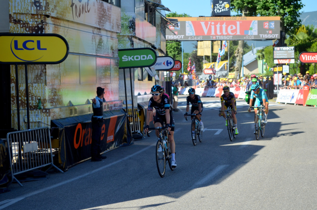 Eine Gruppe von Radfahrern in Helmen und Schutzbrillen fährt eine Straße mit Absperrungen, Schildern und Tafeln mit Text hinunter, während eine Person auf der linken Seite der Straße steht.