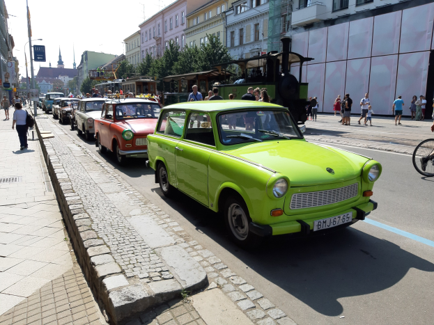 Grünes Auto fährt auf einer Straße mit hohen Gebäuden, Fußgängern auf einem Gehweg, einem Radfahrer, Bäumen und einem klaren blauen Himmel im Hintergrund.