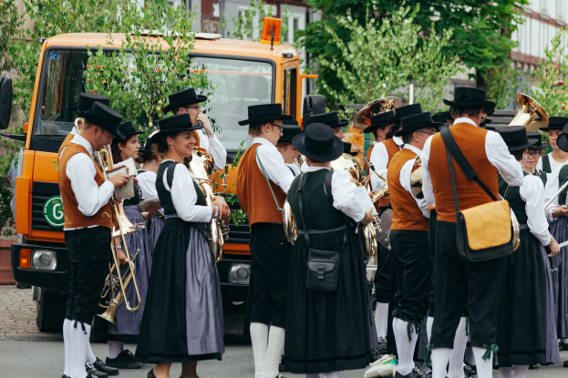 Eine Gruppe von Menschen in traditioneller bayrischer Tracht, die Instrumente spielen und Taschen tragen, mit Bäumen und Gebäuden im Hintergrund und einem geparkten Fahrzeug am Straßenrand.