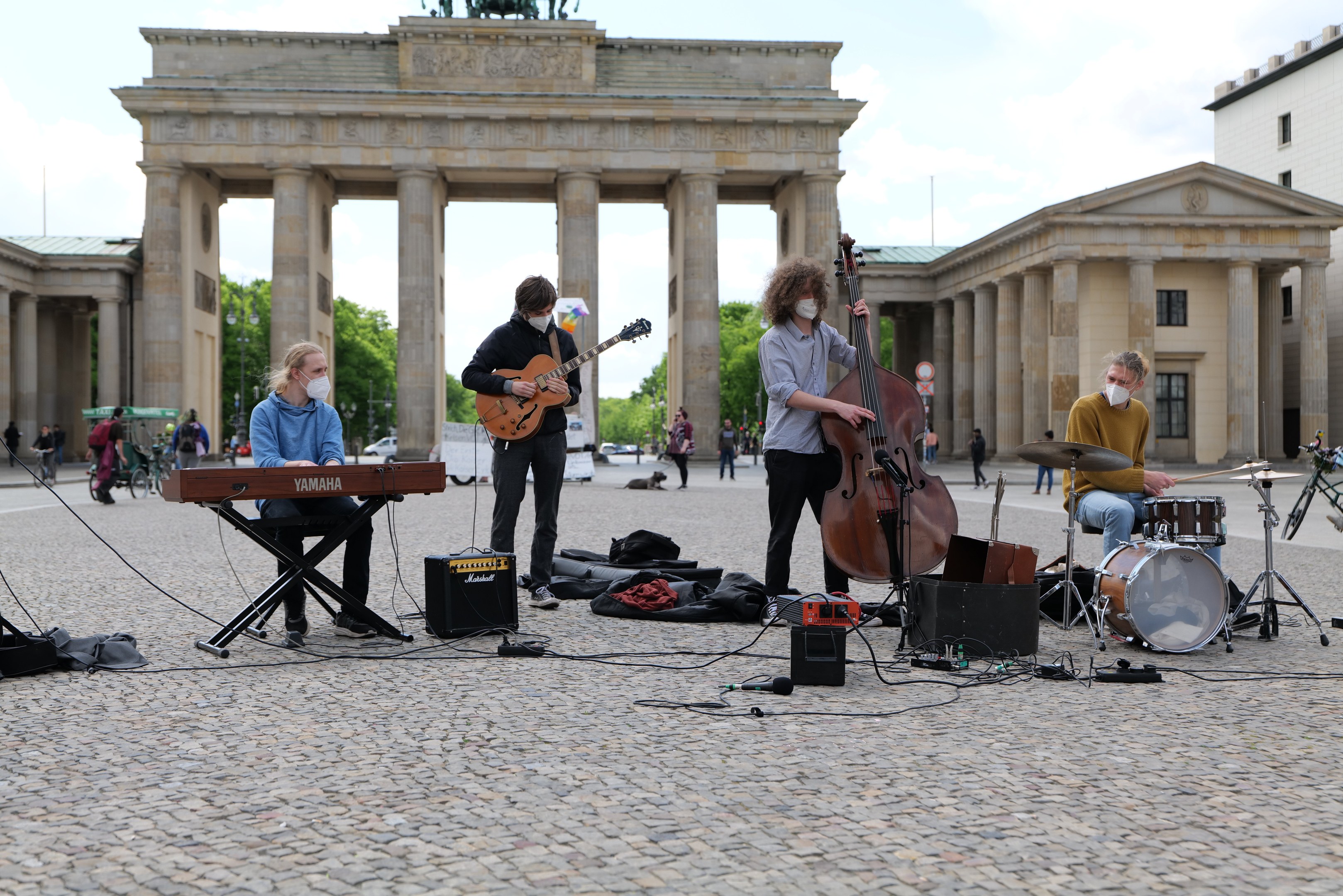 Gruppe von Menschen, die draußen Musik machen, mit einem Gitarristen im Sitzen, einem anderen Musiker und einer Person mit Maske vor dem Brandenburger Tor, mit verstreuter Ausrüstung und städtischer Umgebung.
