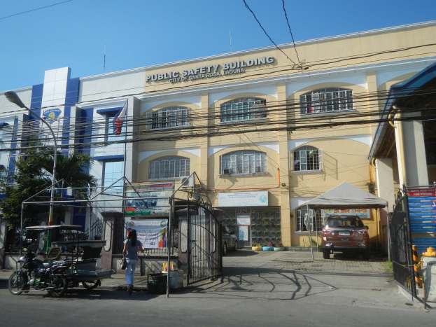 Gebäude umgeben von Bäumen, Straßeninfrastruktur, Fahrzeugen, Fußgängern und einem klaren blauen Himmel.
