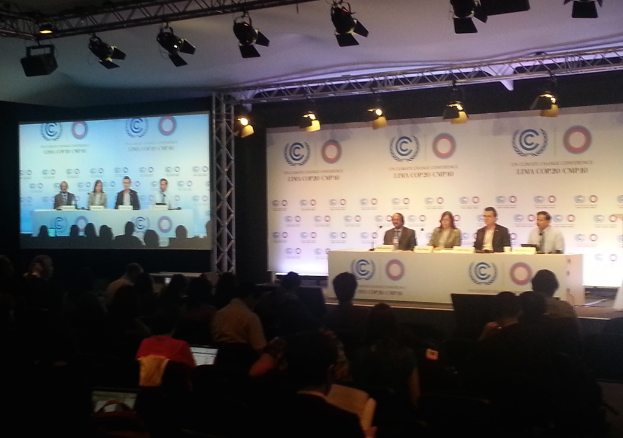 Menschen auf Stühlen auf einer Bühne mit Namensschildern und Mikrofonen auf einem Tisch, Zuschauer im Publikum, eine Projektionsfläche und Beleuchtung an Eisenstangen im Hintergrund.