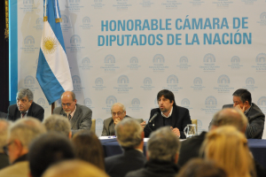 Eine Gruppe von Menschen sitzt an Tischen mit einem Krug, Papieren und einem Stift, während eine Person in der Mitte in ein Mikrofon spricht, mit einer weißen und blauen Flagge und einem Hintergrund, auf dem "ehrenwerte camara de" steht.