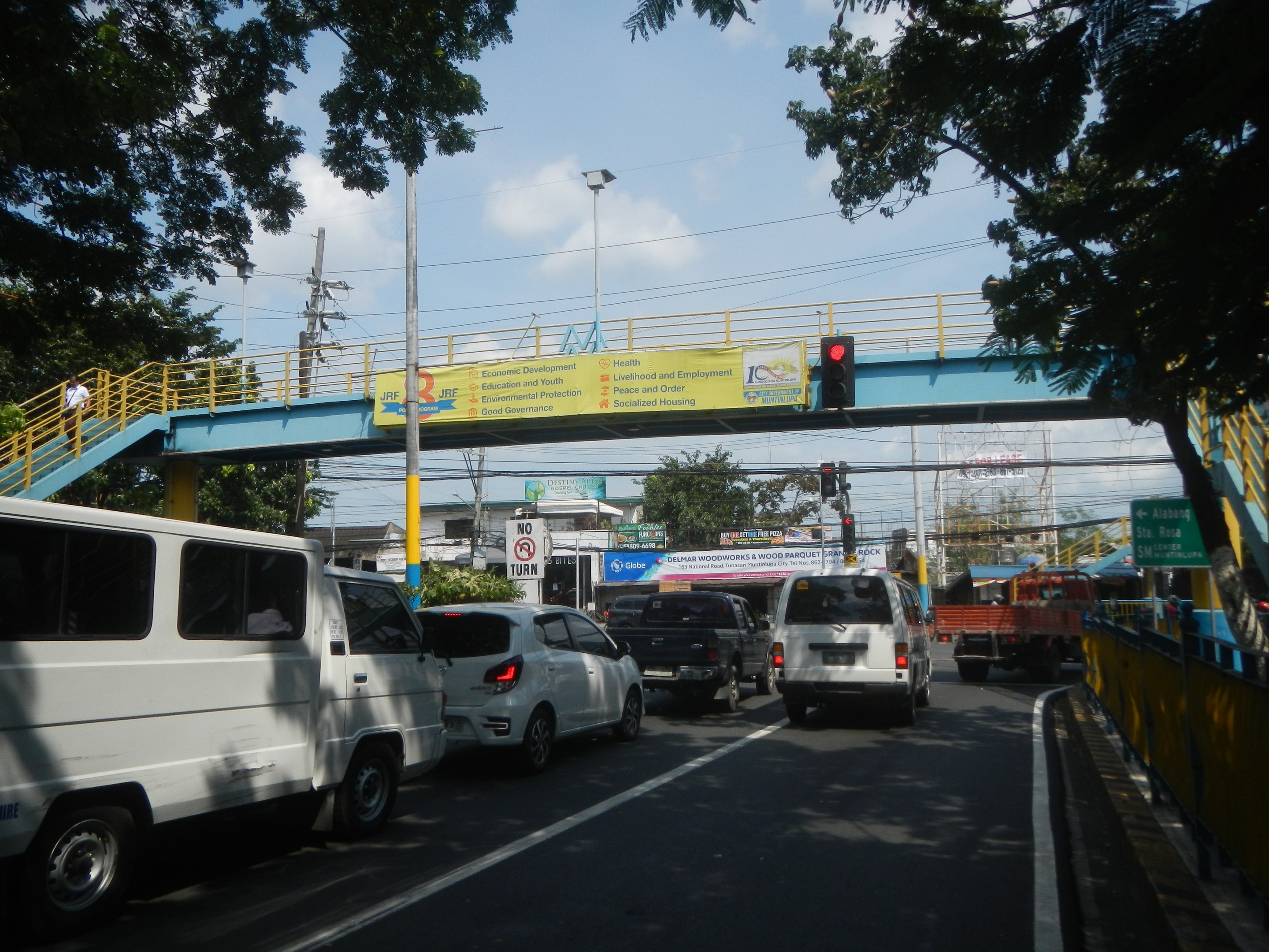 Belebte Straße mit Fahrzeugen, eine Brücke mit Geländer und Treppen, Laternen, Verkehrsampeln, Informationsschilder, Bäume, Gebäude und ein bewölkter Himmel.