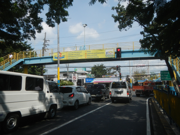 Belebte Straße mit Fahrzeugen, eine Brücke mit Geländer und Treppen, Laternen, Verkehrsampeln, Informationsschilder, Bäume, Gebäude und ein bewölkter Himmel.