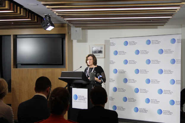 Eine Frau steht auf einer Bühne und spricht in ein Mikrofon, vor ihr sitzen Zuschauer, an der Wand hinter ihr hängt ein Banner, ein Fernseher steht an der Holzwand, und an der Decke leuchten Lampen.