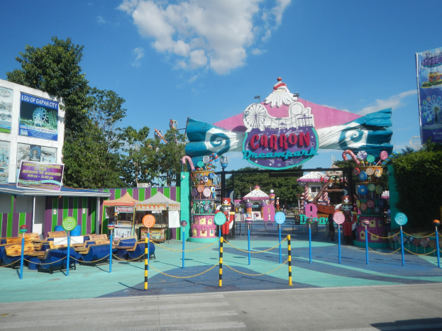 Kirmesfahrgeschäft am Eingang eines Parks, mit Straße, Umzäunung, Buden, Bannern, Bäumen und bewölktem Himmel im Hintergrund.