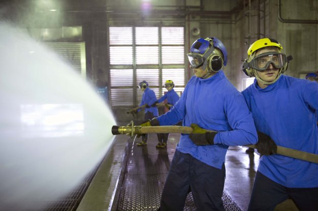 Männer in blauen Hemden und gelben Helmen bei der Arbeit an Maschinen, einer sprüht Wasser auf den Boden in einer Fabrikumgebung mit sichtbaren Rohren, Fenstern und Lampen.