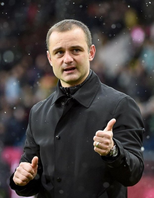 Ein Mann in einer schwarzen Jacke zeigt den Daumen hoch, trägt eine Uhr und hat einen unscharfen Hintergrund, der darauf hindeutet, dass er ein Fußballtrainer ist.