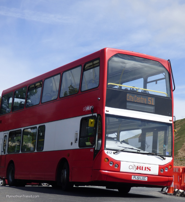 Ein roter Doppeldeckerbus mit der Aufschrift "Stadtbus" fährt auf einer Straße mit einem Verkehrskegel in der Nähe und einem Hügel im Hintergrund.