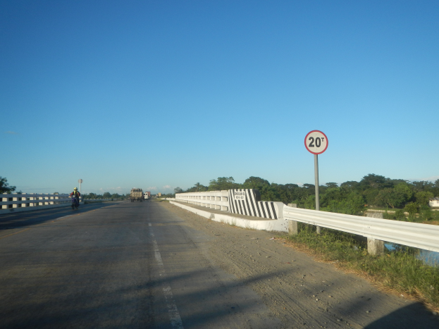 Eine Fahrbahn mit Fahrzeugen, ein Tempolimitschild, Geländer, Gras, Bäume und einen klaren blauen Himmel.