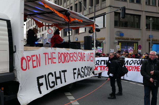 Eine Gruppe von Menschen steht vor einem geparkten Lkw mit einem Transparent mit der Aufschrift "Öffnet die Grenzen, kämpft gegen Faschismus", haltend Schilder und Transparente, mit Gebäuden, Laternenmasten und Verkehrszeichen im Hintergrund.