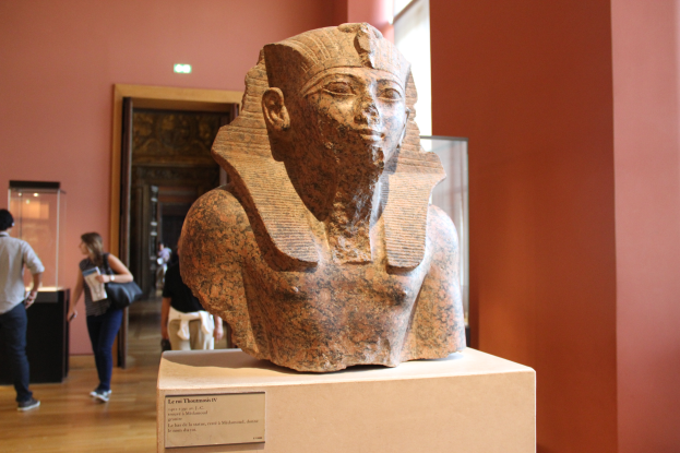 Ein Büste eines ägyptischen Pharaos wird in einem Museum mit einer Texttafel, einer Gruppe von Menschen, einer Frau mit einer Tasche, einer Tür, einer Wand und einer Decke mit Lampen ausgestellt.