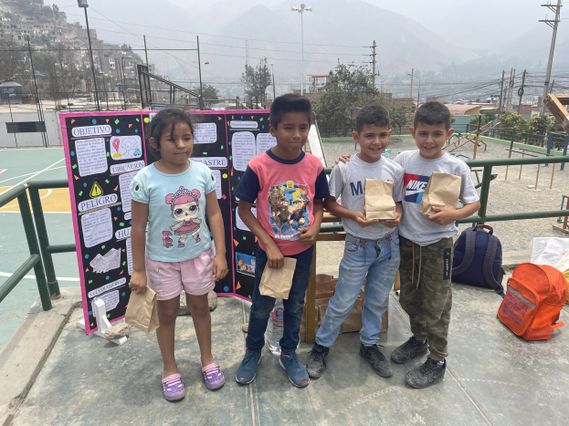 Gruppe von Kindern auf einem Basketballfeld mit Papiertüten in den Händen, mit Tüten und Gegenständen rechts und einer Tafel mit Text links, vor einem Hintergrund aus Zaun, Pfosten, Laternen, Strommasten, Bäumen, Gebäuden, Hügeln und Himmel.