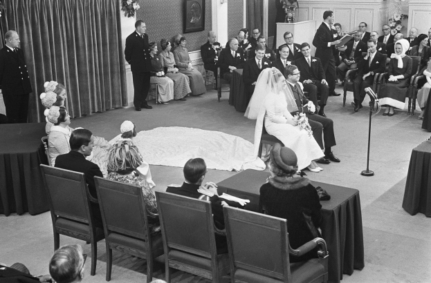 Schwarzes und weißes Foto einer Hochzeitszeremonie mit sitzenden und stehenden Gästen, einem Mann mit Mikrofon, einer Frau mit Blumenstrauß, einem Podium mit Mikrofon auf der rechten Seite, Blumengestecken und einem Vorhang auf der linken Seite, einem gerahmten Foto an der Wand und einer Tür im Hintergrund.