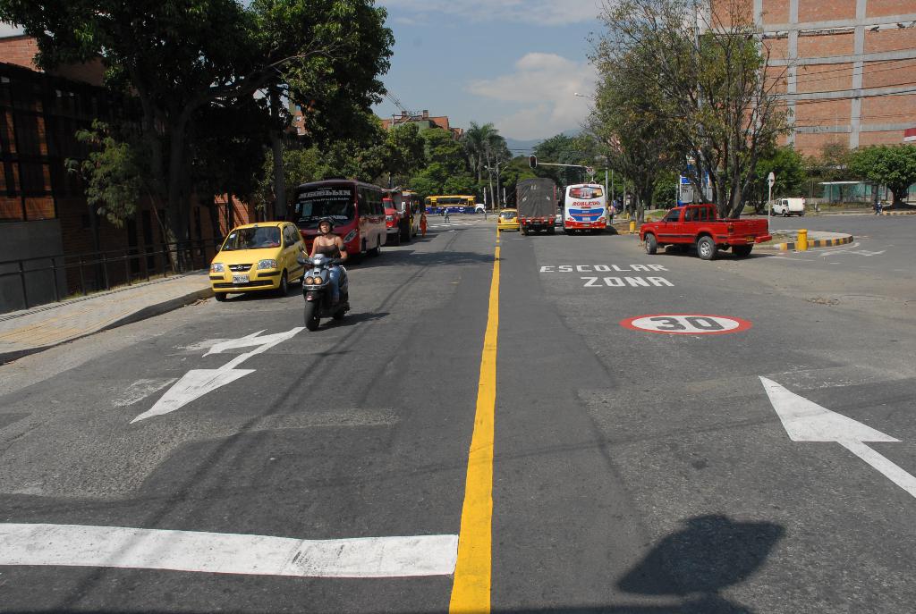 Eine Straßenszene mit Gebäuden, Bäumen, fahrenden Fahrzeugen, Schildern, Verkehrsampeln, einem blauen bewölkten Himmel und Warnschildern auf der Straße.