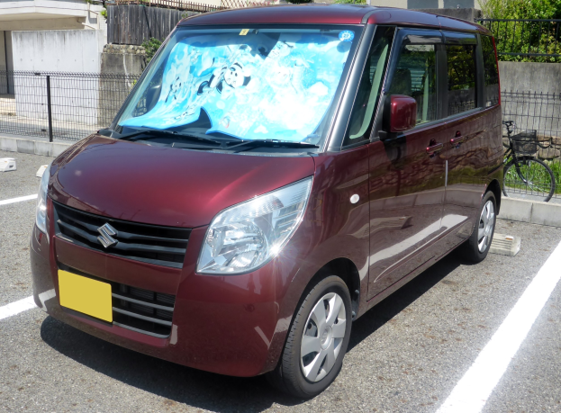 Ein roter Minivan auf einem Parkplatz neben einem Gebäude geparkt, mit einem Fahrrad auf der rechten Seite und einem Geländer im Hintergrund sowie einigen Pflanzen und Bäumen.