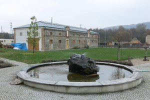 Ein Brunnen mit einem großen Felsen in der Mitte, umgeben von grünem Gras und Bäumen, mit einem Gebäude, einem geparkten Fahrzeug und Strommasten mit Drähten im Hintergrund, aufgenommen in Tschechien im Jahr 2015.