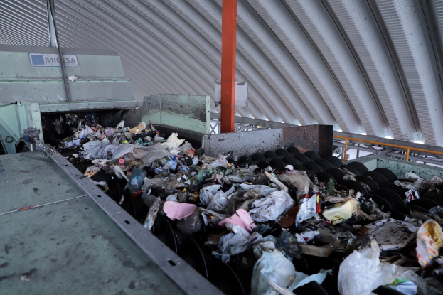 Großer Müllberg auf einem Förderband in einer Recyclinganlage mit Maschine und Metalldach.