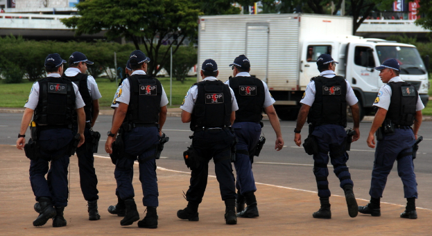 Eine Gruppe von Beamten in Uniform, darunter MÃ¼tzen, Hemden, kugelsichere Westen, Uhren, Hosen und Schuhe, die auf einer StraÃe mit einem geparkten Lkw auf der rechten Seite nÃ¤her an einem Zaun, GebÃ¤uden, BÃ¤umen, Pflanzen, Masten und Bannern im Hintergrund und einer BrÃ¼cke im oberen rechten Eck zu sehen sind.