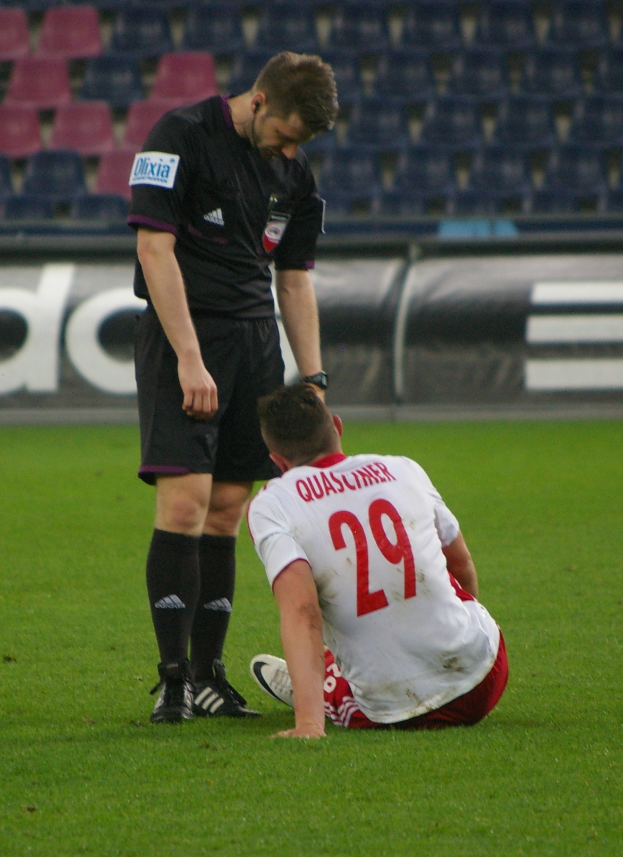 Ein Füßballspieler sitzt neben einem Schiedsrichter auf dem Boden in einem Stadion, beide tragen Sportkleidung, mit Plakaten und Stühlen im Hintergrund.