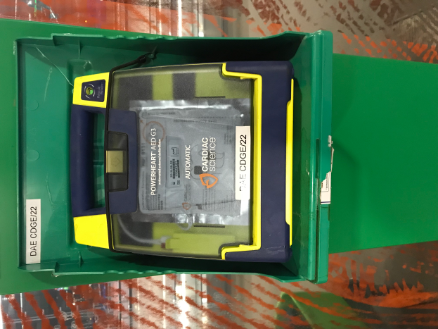 Ein grüner Kasten mit einem gelben und blauen Defibrillator-Gerät darin, das auf einem Tisch steht.